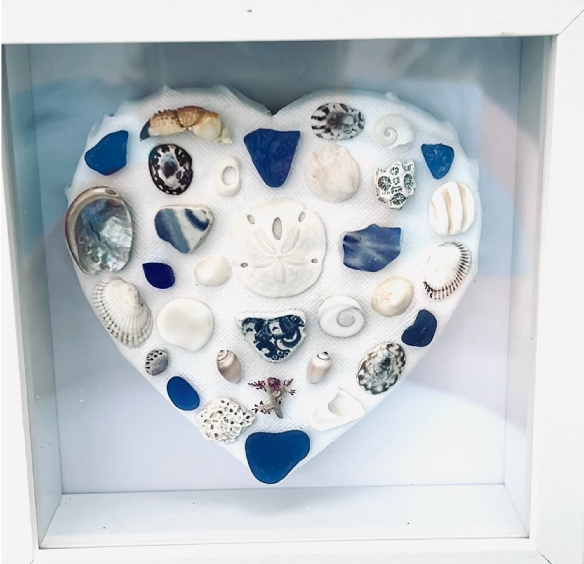 Heart-shaped display of seashells and beach stones in a white frame